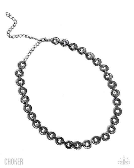 A dark gunmetal silver choker necklace with textured circular links, displayed on a clean white background with a 'CHOKER' text overlay on the bottom left.