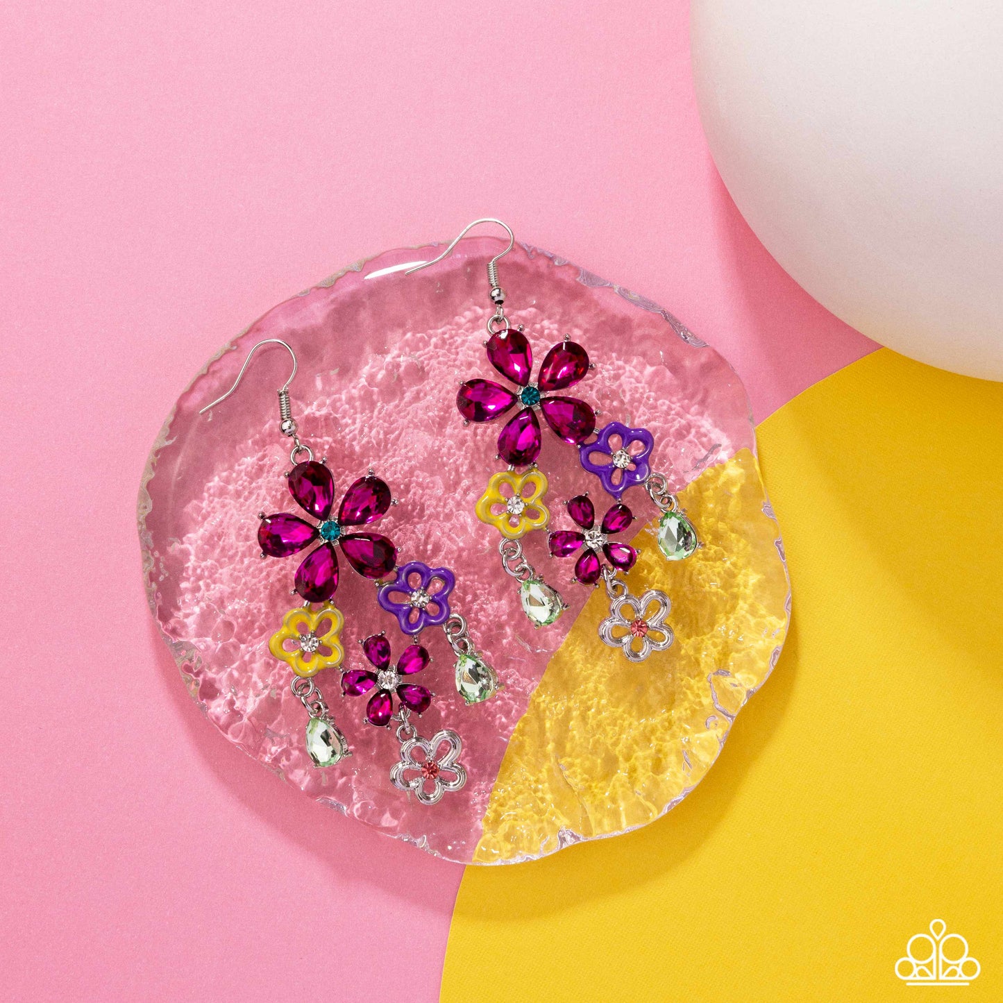 Colorful flower-shaped earrings on a pink and yellow heart-shaped stand with a pink background.
