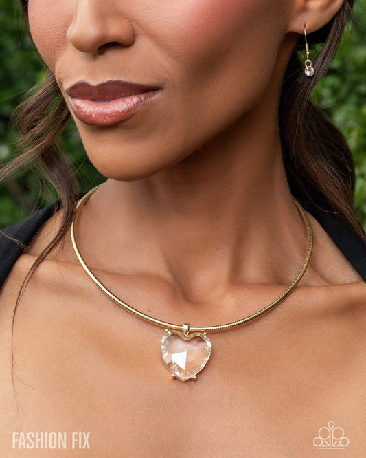 Close-up of a woman wearing a gold necklace with a heart-shaped pendant against a blurred green background.