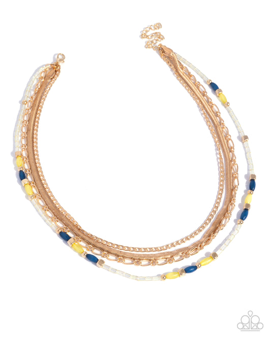 A multi-layered gold chain necklace with white beads and blue cylindrical accents, displayed on a clean white background.