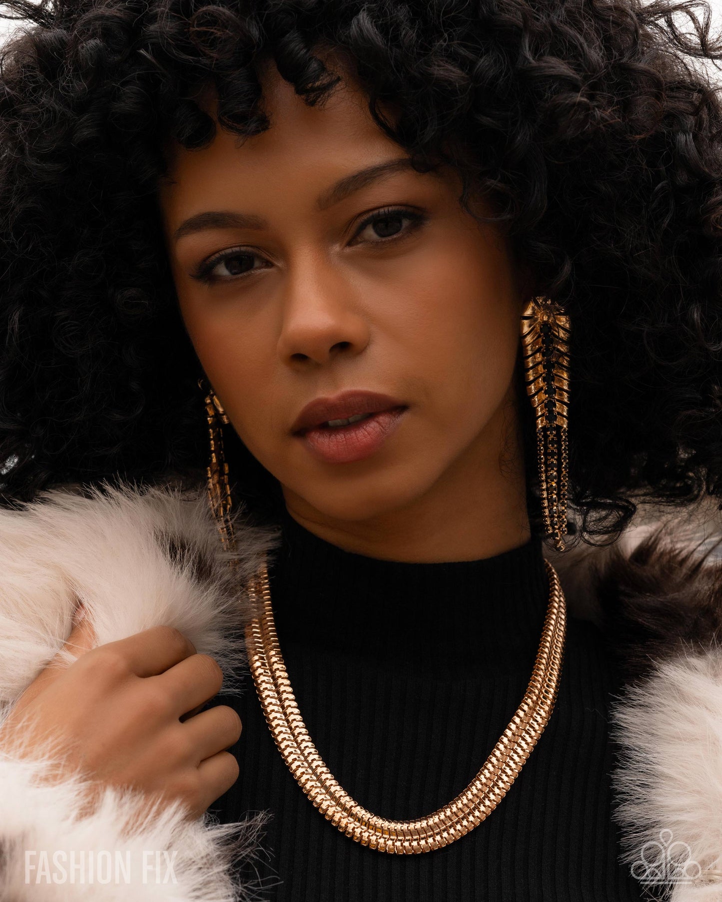 Woman wearing a gold necklace and earrings with a fur coat, posing against a neutral background.