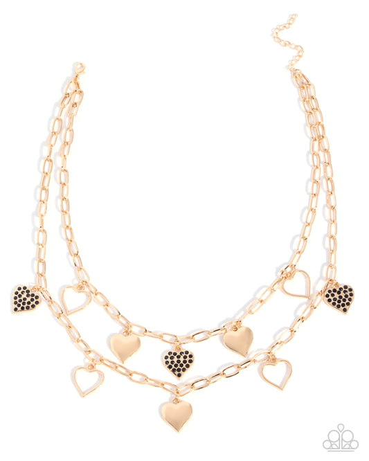 A double-layered gold-tone chain necklace with heart charms, featuring some heart charms encrusted with small black stones, displayed on a clean white background.