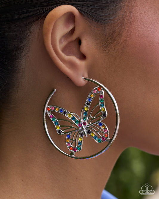 A close-up side profile of a person wearing a large silver hoop earring with a butterfly design set with multicolored rhinestones.