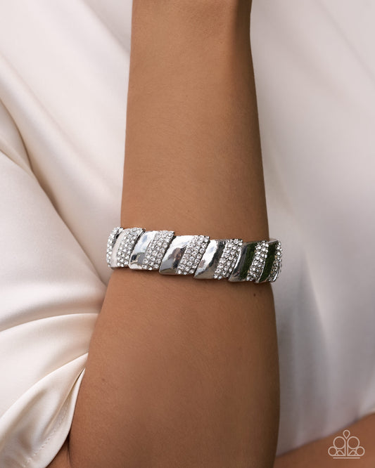 A close-up of a person's wrist wearing a chunky silver cuff bracelet embellished with rows of clear crystals, set against a soft cream-colored satin background.