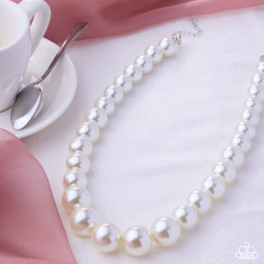 Pearl necklace on a pink surface with a cup and spoon in the background