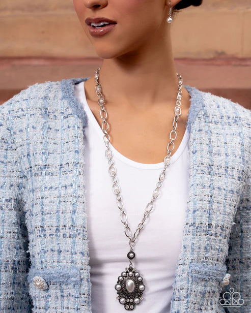 Woman wearing a long silver necklace with a floral pendant, light blue cardigan, and white top.