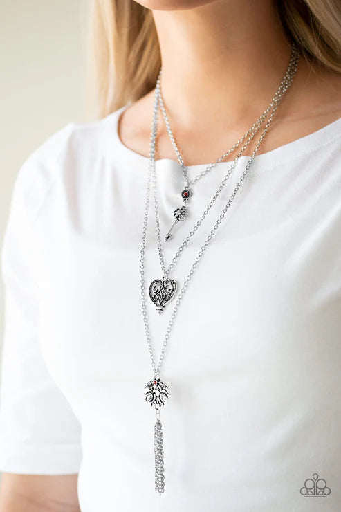 Layered silver necklaces with heart and tassel pendants on a white shirt.