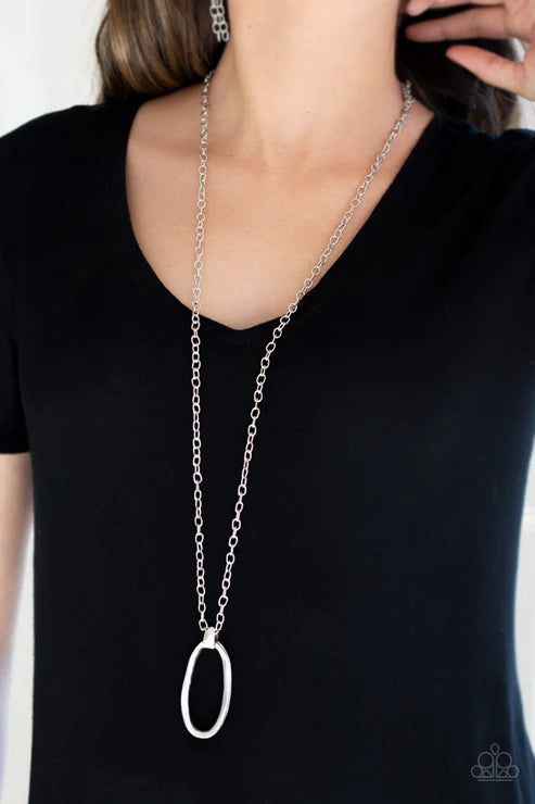 Person wearing a long silver chain necklace with an oval pendant on a plain background