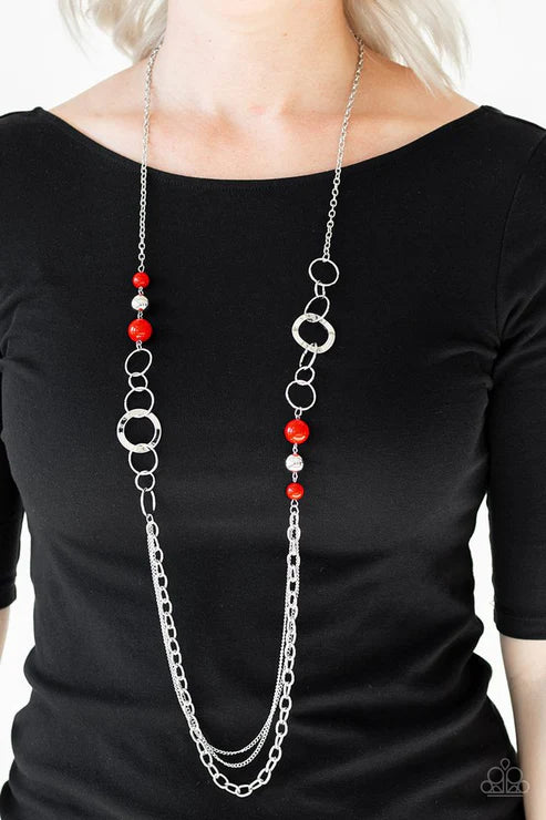 Long silver chain necklace with red beads worn by a person on a plain background