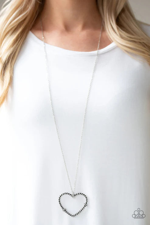 Long necklace with a heart-shaped pendant worn by a person in a white outfit.