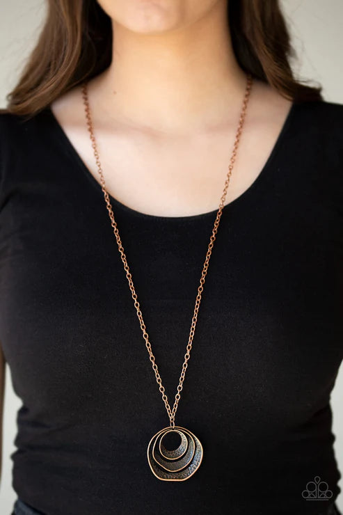 Person wearing a long necklace with a circular pendant on a plain background