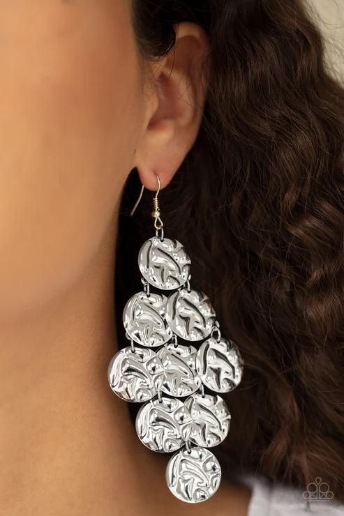 A close-up of a person's ear wearing a large silver dangle earring made of stacked textured circular discs.