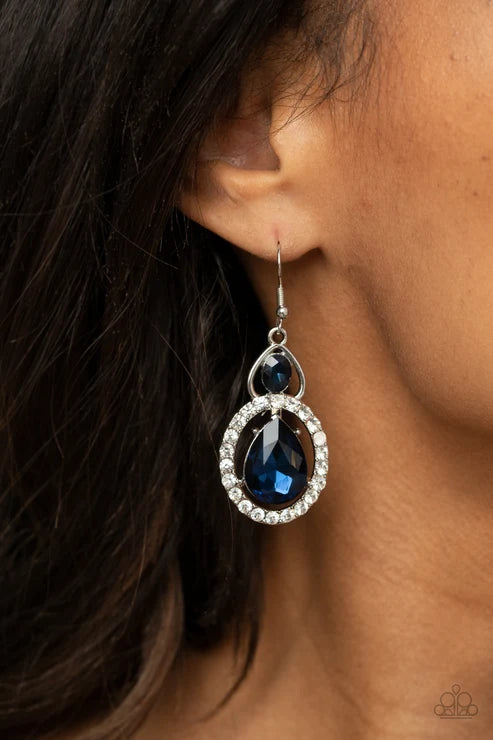 A close-up of a model's ear wearing a silver-toned dangle earring with a large faceted dark blue teardrop gemstone surrounded by a halo of small clear crystals, with a smaller dark blue stone above.