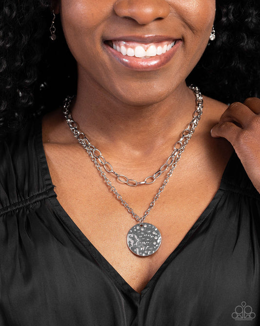 Woman wearing a silver necklace with a round pendant against a dark background