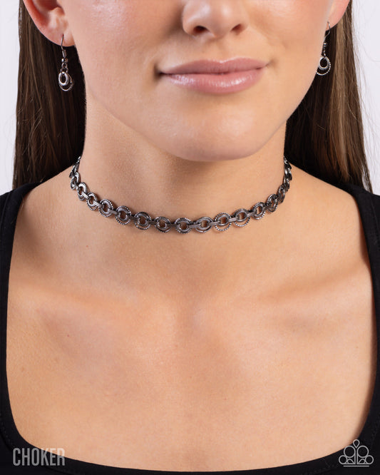 A close-up of a woman wearing a dark metallic chain choker necklace and matching dangling earrings against a light gray background.