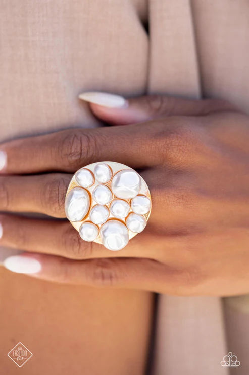 Close-up of a hand wearing a large, round, pearl-studded ring against a neutral background.