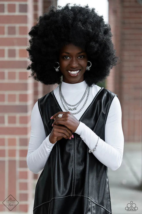 Woman wearing a black leather vest over a white long-sleeve shirt, standing against a brick wall.