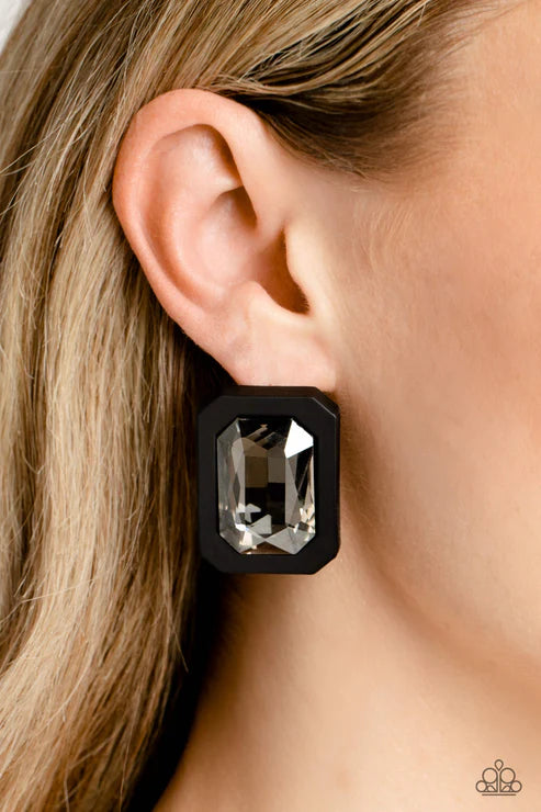 A close-up of a model's ear wearing a black geometric stud earring with a faceted clear gemstone. The model has blonde hair, and a small Paparazzi logo watermark is visible in the bottom right corner.