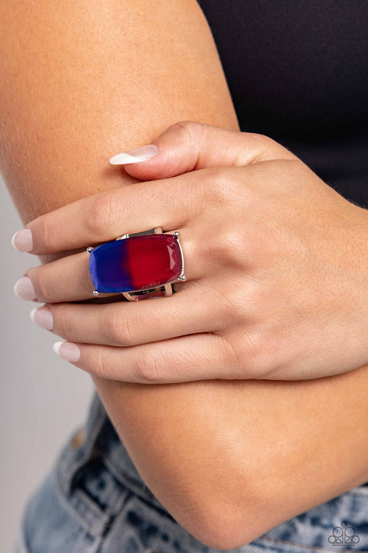 Close-up of a hand wearing a red and blue ring on a neutral background