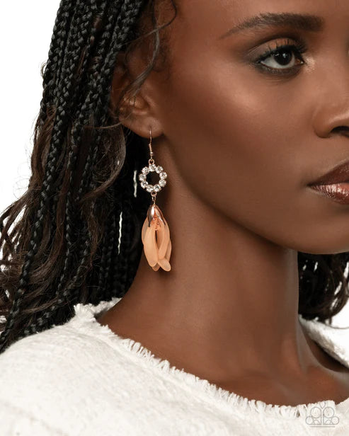 A close-up of a woman wearing a dangling earring with a silver-tone circular crystal halo and a peach-colored leaf-shaped charm, against a white background.