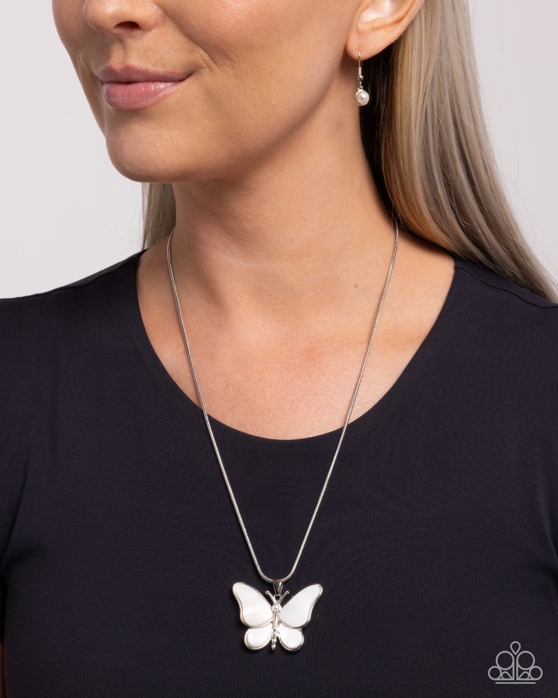 A close-up of a woman wearing a silver butterfly pendant necklace with a white inlay, paired with a silver drop earring. The woman is wearing a black top against a light gray background.
