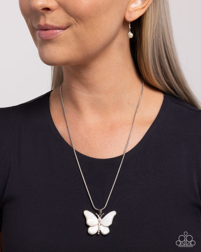 A close-up of a woman wearing a silver butterfly pendant necklace with a white inlay, paired with a silver drop earring. The woman is wearing a black top against a light gray background.