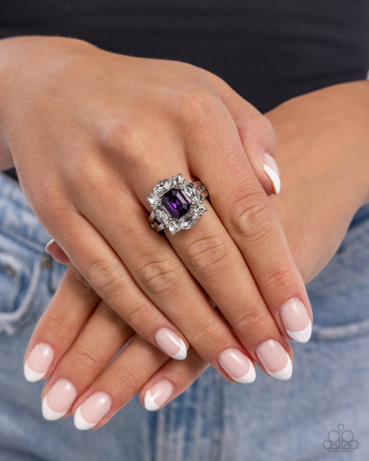 Close-up of a hand wearing an ornate ring with a purple gemstone, set against a blurred denim background.