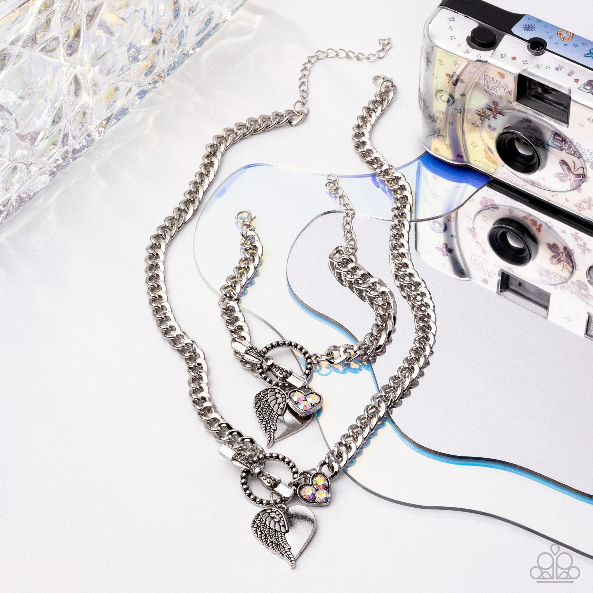 Silver chain necklace with heart and wing charms on a white surface with a vintage camera.