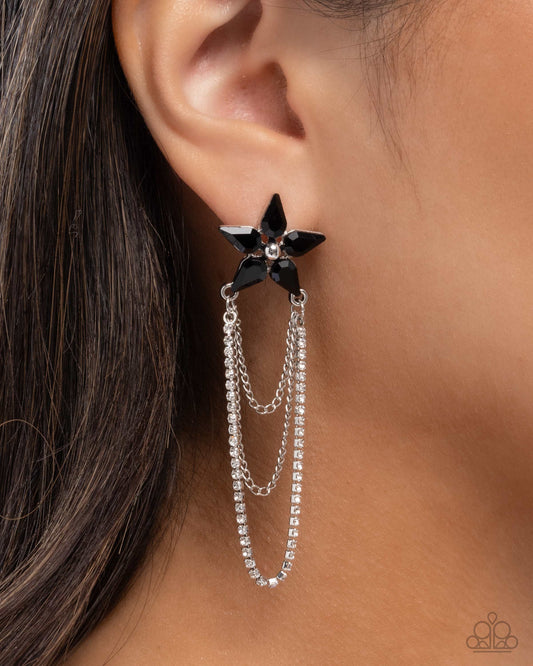 A close-up of a model's ear wearing a Paparazzi brand black star-shaped stud earring with dangling silver chains set with clear rhinestones.