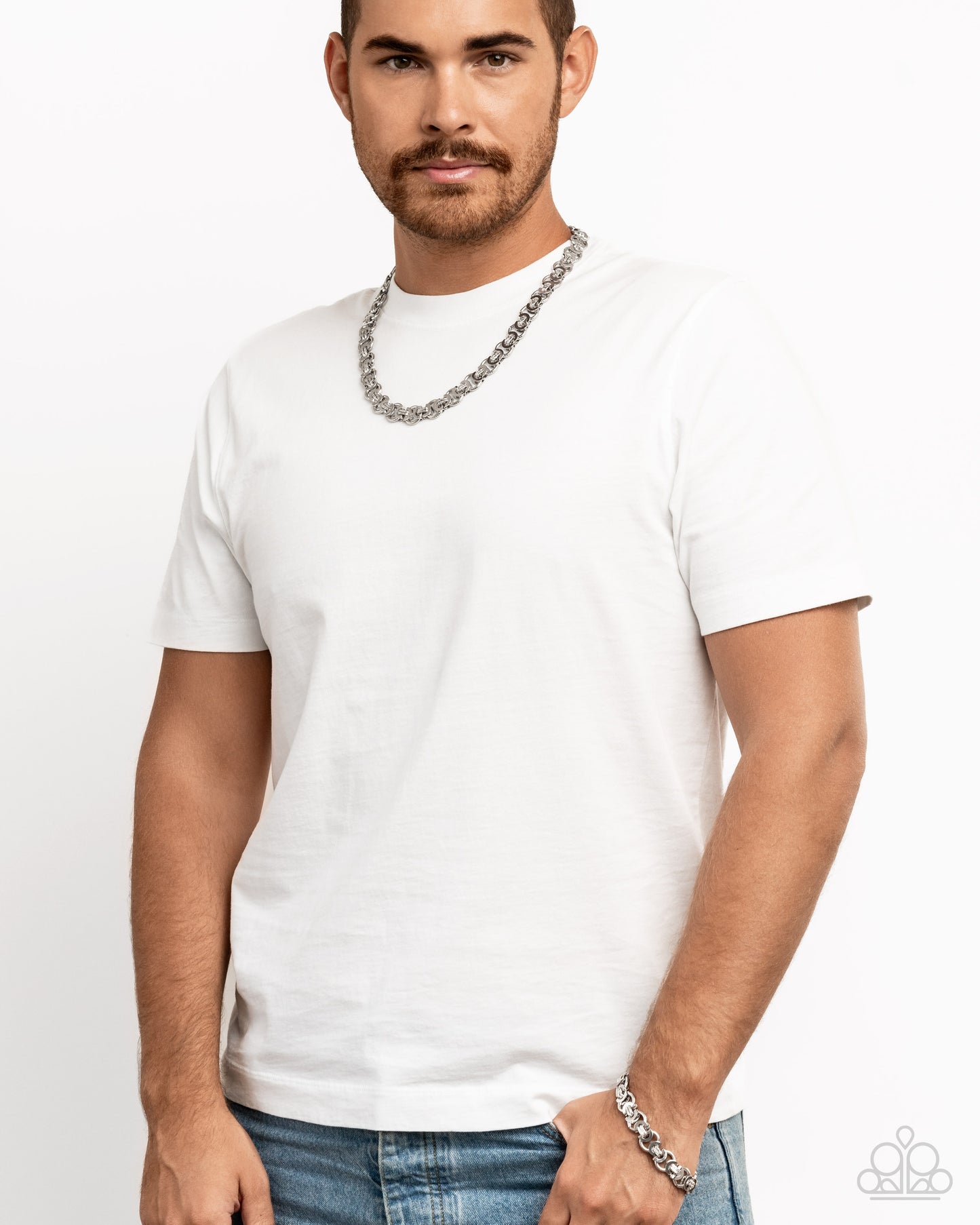 Man wearing a white t-shirt and blue jeans with a silver chain necklace on a plain background