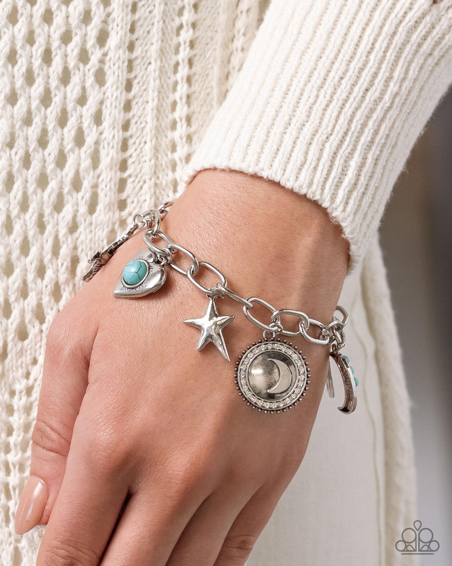 A close-up of a silver charm bracelet with a turquoise stone, star, moon, and crescent moon charms, worn on a person's wrist over a cream knit sweater.