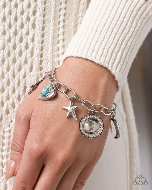 A close-up of a silver charm bracelet with a turquoise stone, star, moon, and crescent moon charms, worn on a person's wrist over a cream knit sweater.
