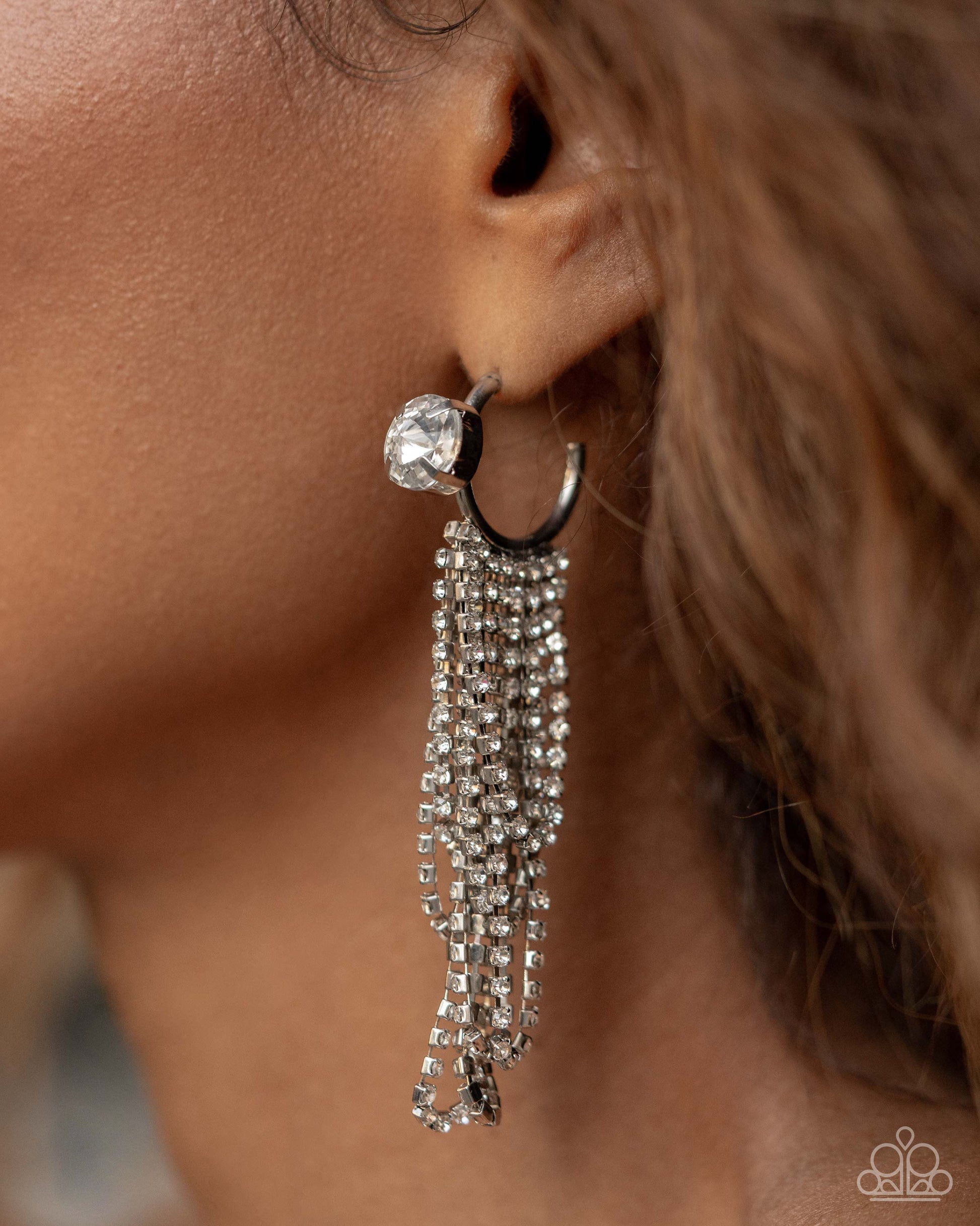A close-up of a model's ear wearing a silver-toned huggie hoop earring adorned with a large clear crystal and multiple dangling chain strands of clear crystals.