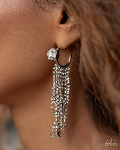 A close-up of a model's ear wearing a silver-toned huggie hoop earring adorned with a large clear crystal and multiple dangling chain strands of clear crystals.