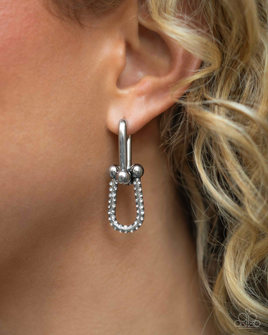 A close-up of a model's ear wearing a silver hoop earring with a dangling safety-pin-inspired design set with small clear stones.