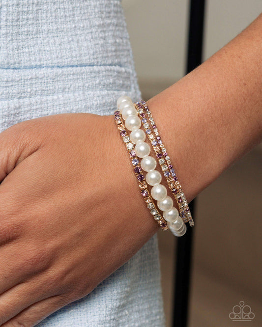 Three stacked bracelets on a person's wrist, featuring a strand of white faux pearls and two rose gold bands set with clear rhinestones and small purple crystals, worn with a light blue textured top.