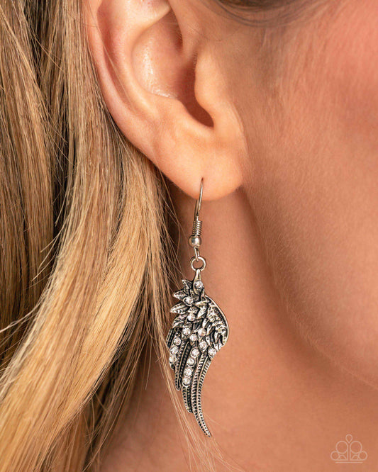 A close-up of a model's ear wearing a silver wing-shaped dangle earring adorned with clear rhinestones. The background is filled with the model's blonde hair.