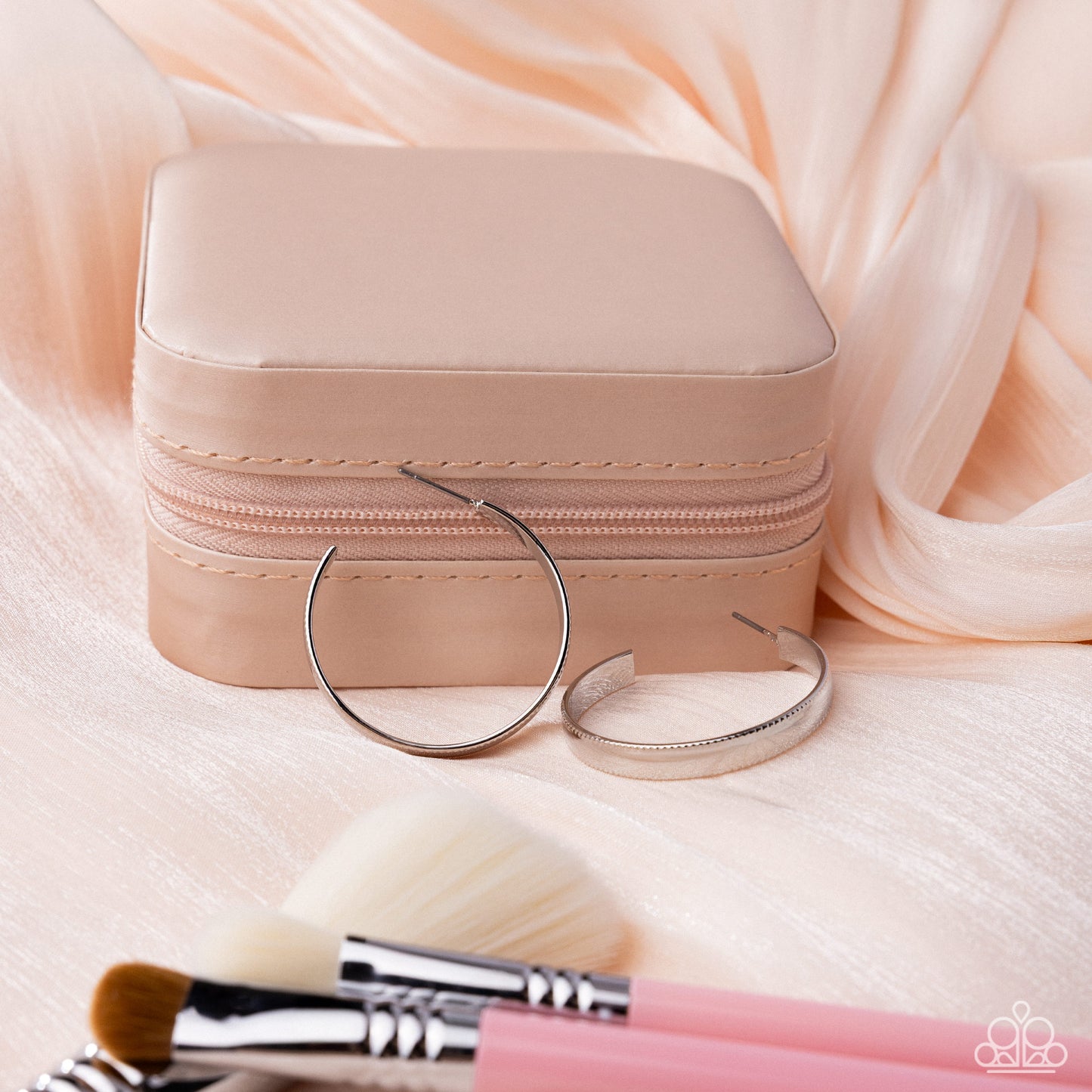 Small beige jewelry box with silver hoop earrings and makeup brushes on a soft pink fabric background