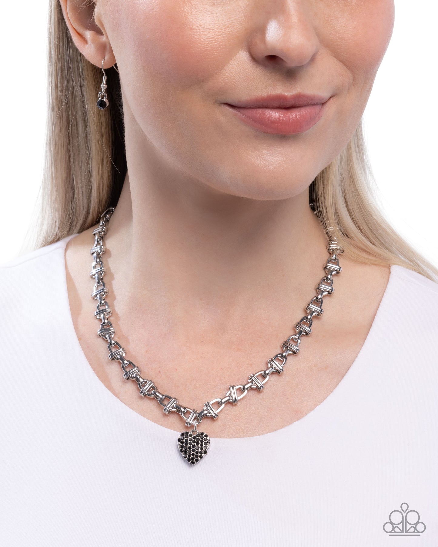 A close-up of a woman wearing a silver-toned necklace with a heart-shaped pendant and matching drop earrings, shown against a plain white background.