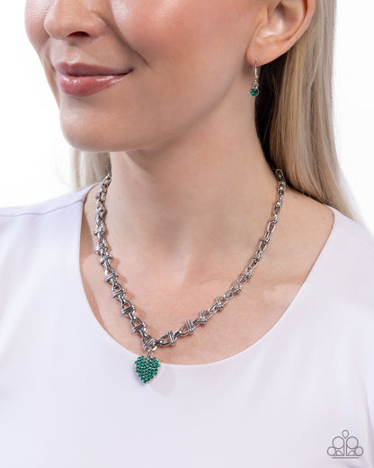A close-up of a woman wearing a silver chain necklace with a green heart-shaped crystal pendant, along with matching green and silver drop earrings, against a white background.