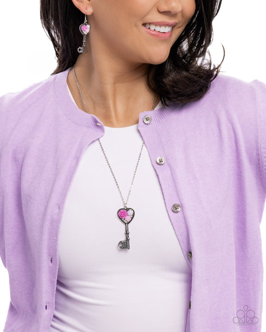 Woman wearing a purple cardigan with a pink heart key pendant necklace.