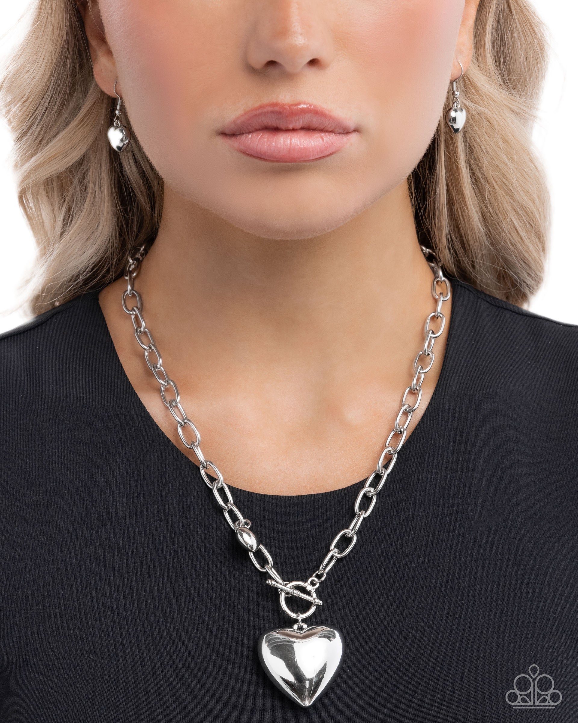 Person wearing a silver chain necklace with a heart pendant on a plain background