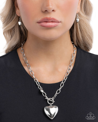 Person wearing a silver chain necklace with a heart pendant on a plain background