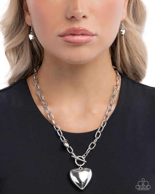 Person wearing a silver chain necklace with a heart pendant on a plain background