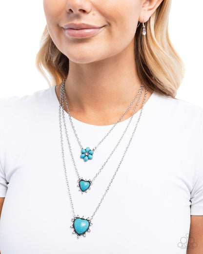 A close-up of a woman wearing a layered silver necklace set with turquoise blue pendants, including a flower-shaped charm, a heart pendant, and a larger heart pendant, paired with a white t-shirt.