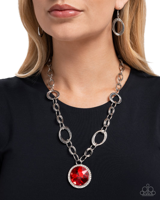 Person wearing a silver chain necklace with a red gemstone pendant on a black background
