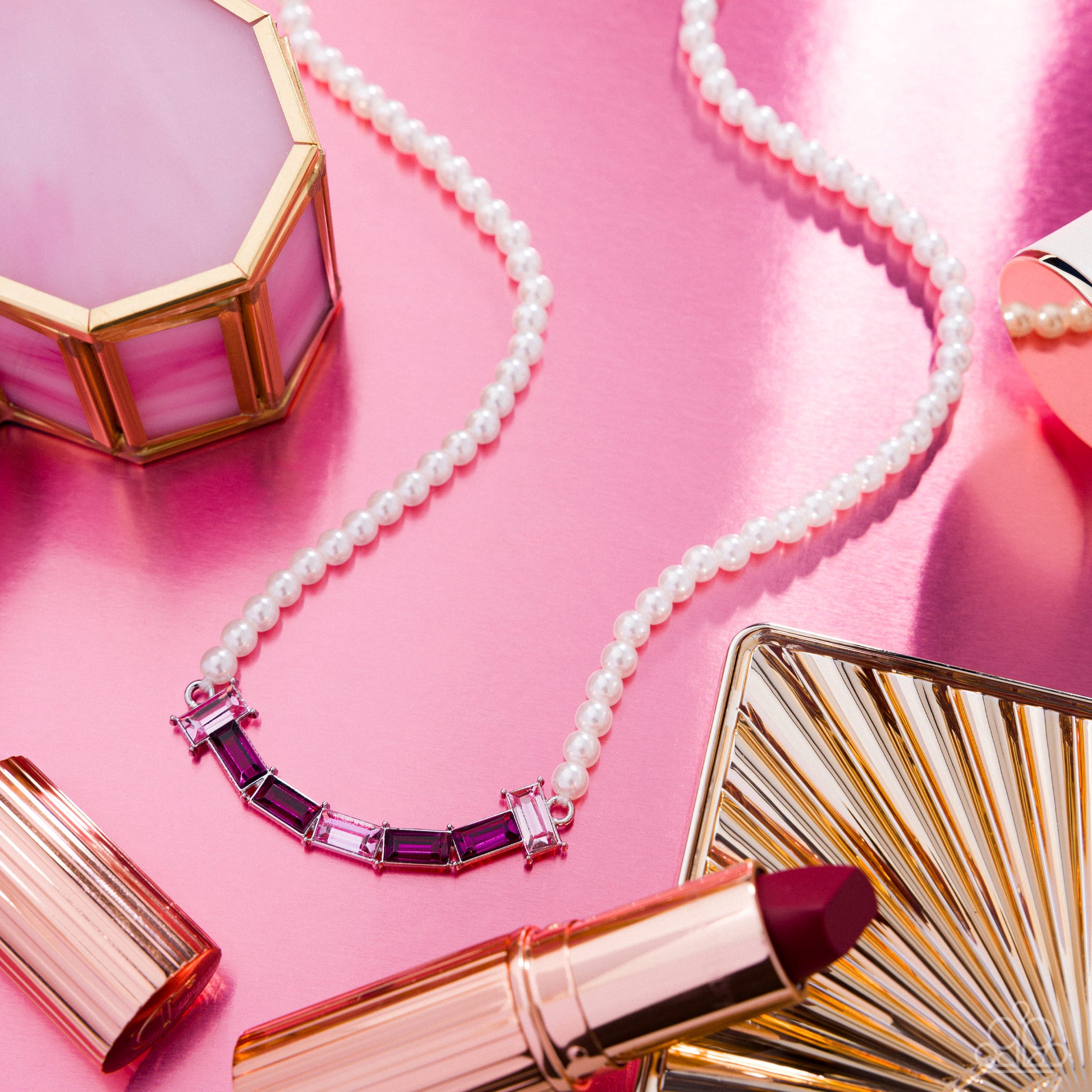 Makeup items including a lipstick, pearls, and a compact mirror on a pink background