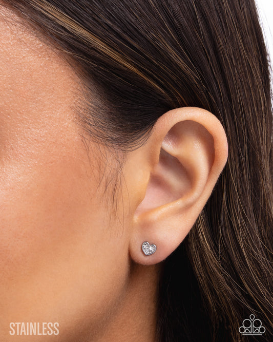 Close-up of an ear wearing a silver heart-shaped stud earring with 'Stainless' text below.