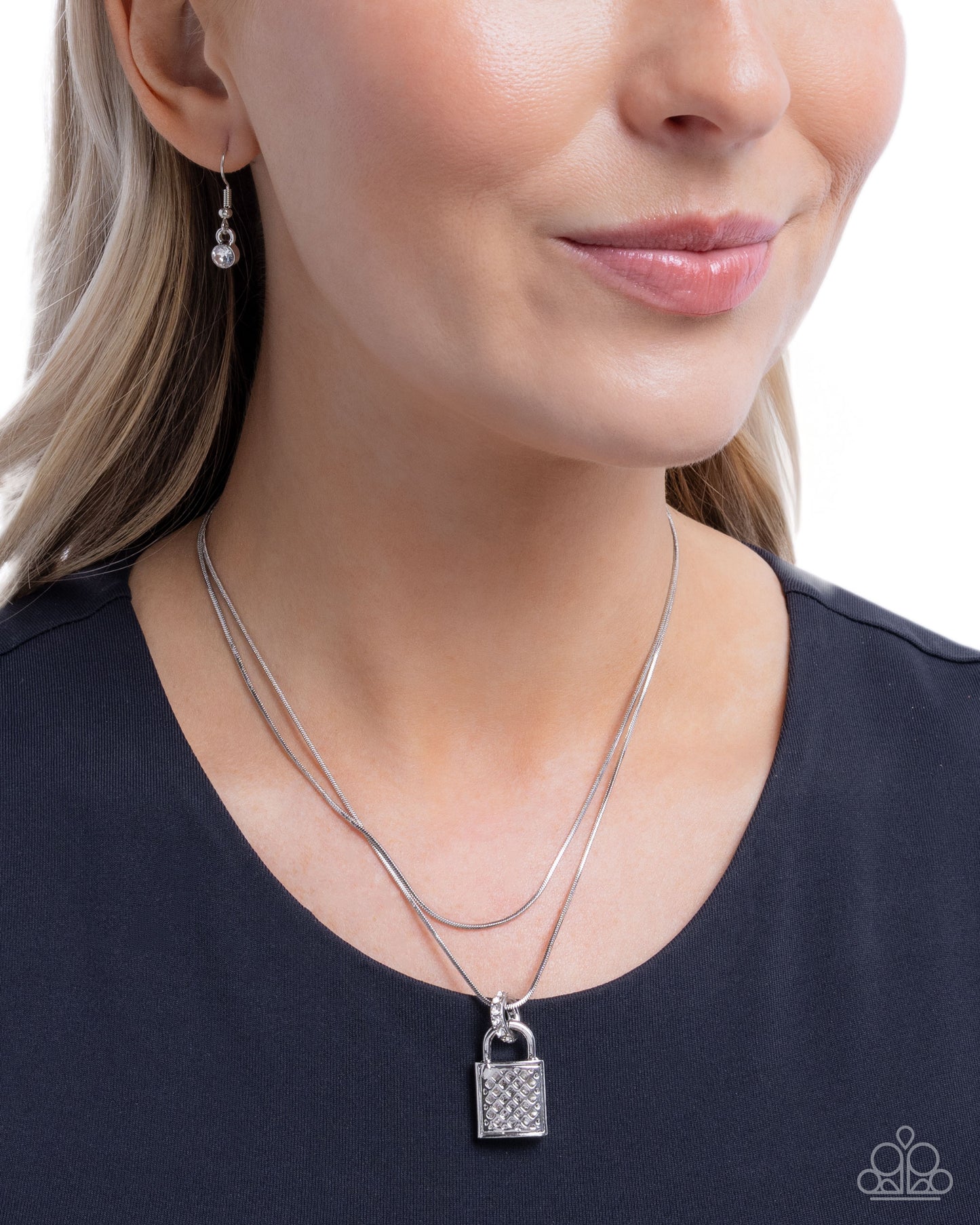 A close-up of a woman wearing a silver double-strand necklace with a small padlock pendant, along with a matching drop earring, against a white background.