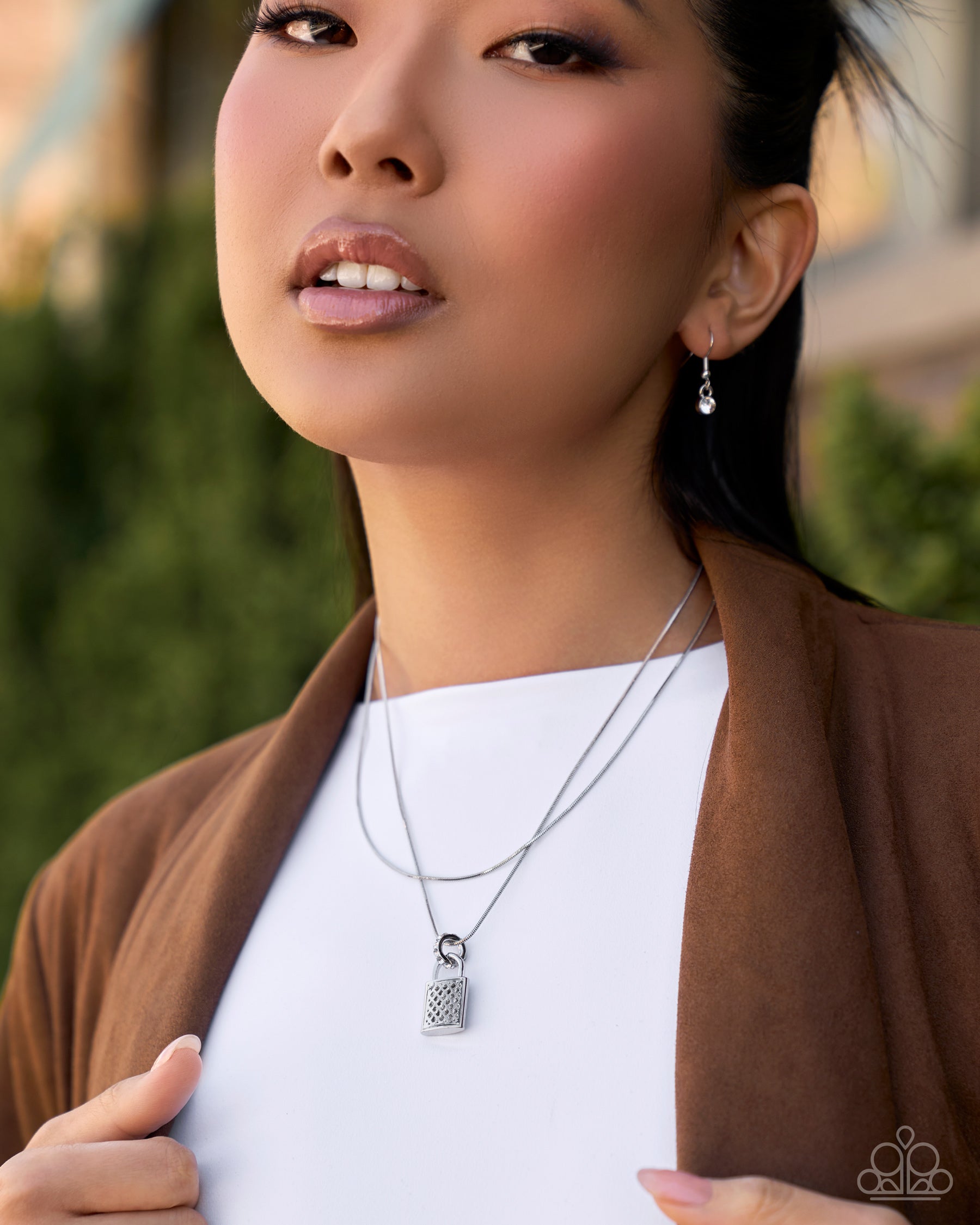 A close-up lifestyle shot of a woman wearing a silver double-strand necklace with a textured padlock pendant, styled with a white top and brown cardigan.